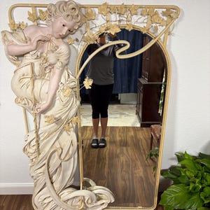 May include: A large, ornate mirror with a white and gold finish. The mirror features a sculpted figure of a woman with flowing hair and a dress. The frame is decorated with leaves and vines.