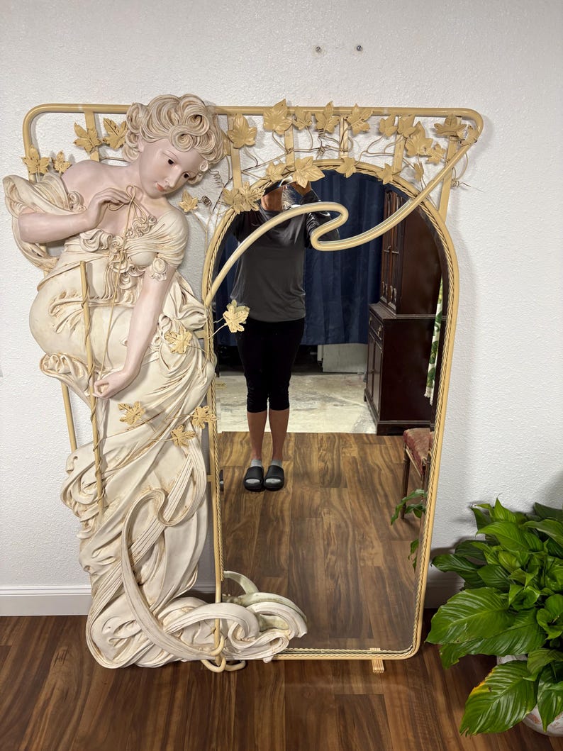 May include: A large, ornate mirror with a white and gold finish. The mirror features a sculpted figure of a woman with flowing hair and a dress. The frame is decorated with leaves and vines.