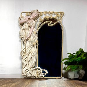 Huge Alphonse Mucha Style Art Nouveau Mirror with a life-size plaster resin statue of a 66” tall young woman in a flowing cream-colored dress. The arched mirror on the right side of the woman is surrounded by a wrought iron frame with vines and flowers along the top.
