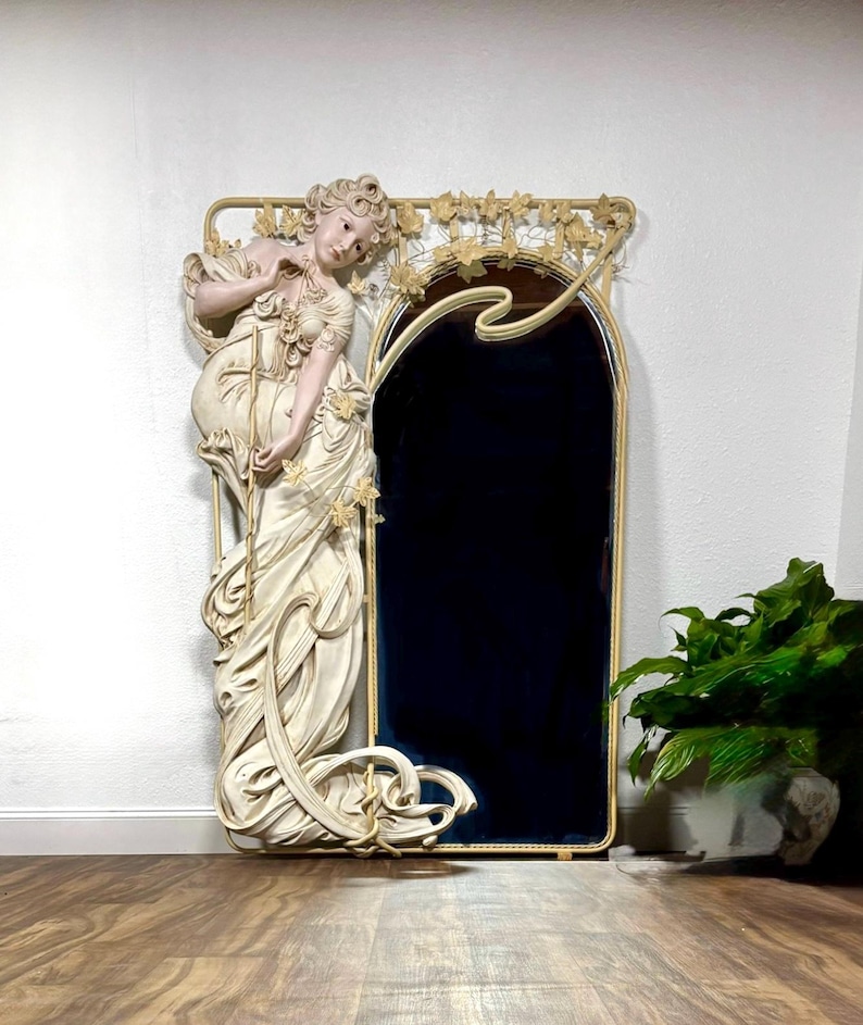 Huge Alphonse Mucha Style Art Nouveau Mirror with a life-size plaster resin statue of a 66” tall young woman in a flowing cream-colored dress. The arched mirror on the right side of the woman is surrounded by a wrought iron frame with vines and flowers along the top.