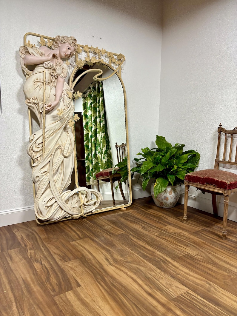 May include: A large, ornate, metal-framed mirror with a white, sculpted figure of a woman standing in a flowing gown. The mirror is leaning against a wall with a light brown wood floor. There is a green plant in a white pot and a wooden chair with a red cushion in the background.