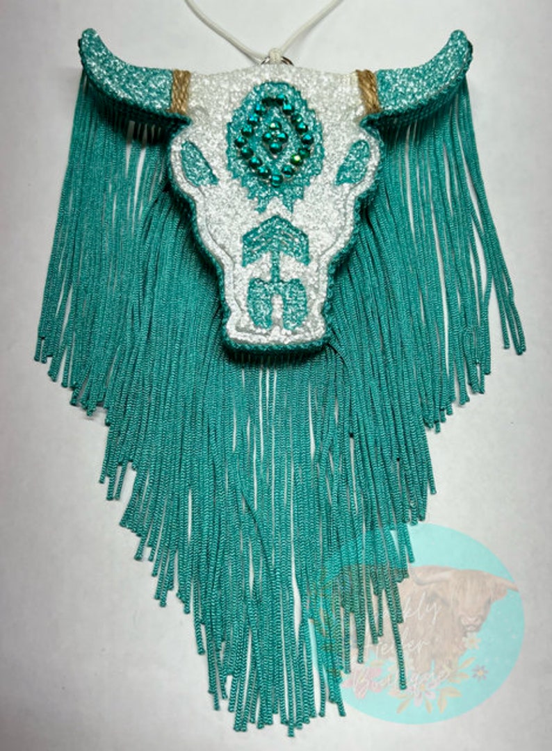 May include: White glitter bull skull with turquoise fringe. The skull has a beaded design on the forehead.