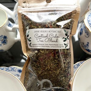May include: A clear plastic bag filled with a tea blend, labeled "Cortisol Calm Tea Blend" with ingredients listed. The bag is set against a backdrop of white teacups and teapots with blue floral patterns, suggesting a tea-related theme.