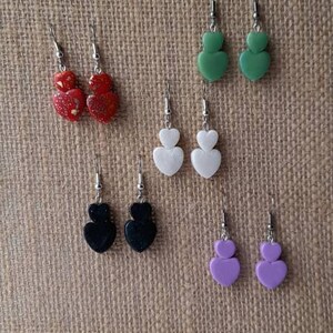 May include: Assortment of double heart shaped earrings in various colours. The earrings are displayed on a burlap background. The colours include green, white, black, and purple.