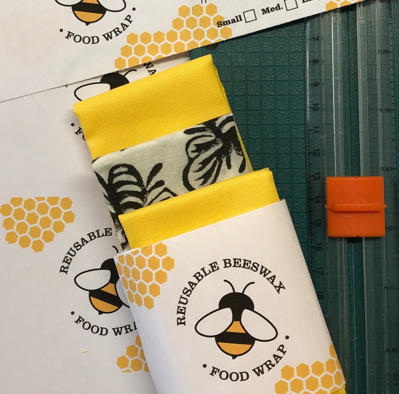 Pode incluir: Um conjunto de tr&ecirc;s embalagens de alimentos reutiliz&aacute;veis de cera de abelha com um design amarelo, branco e preto. As embalagens s&atilde;o embaladas em um envelope branco com uma ilustra&ccedil;&atilde;o de abelha e o texto "Embalagem de cera de abelha reutiliz&aacute;vel".