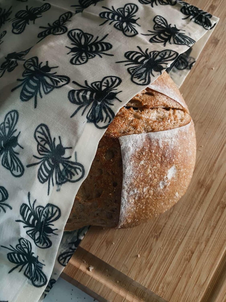 May include: A round loaf of crusty brown sourdough bread partially covered by a cream-colored fabric wrap featuring a repeating pattern of black bees. The bread has a visible score mark across the top and a dusting of flour on its surface.