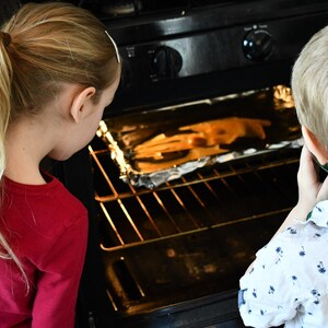 May include: Two children, one with blonde hair in a ponytail wearing a red shirt and the other with blonde hair wearing a white shirt with a blue pattern, look into an oven with a baking sheet inside.