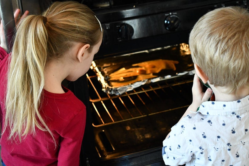 May include: Two children, one with blonde hair in a ponytail wearing a red shirt and the other with blonde hair wearing a white shirt with a blue pattern, look into an oven with a baking sheet inside.