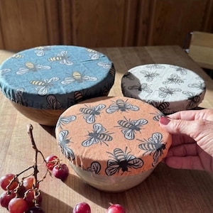 May include: Three bowls with fabric covers featuring bee patterns. The covers are in shades of blue, peach, and white. The bowls are on a wooden table, with a bunch of grapes in the foreground. The fabric covers are reusable.