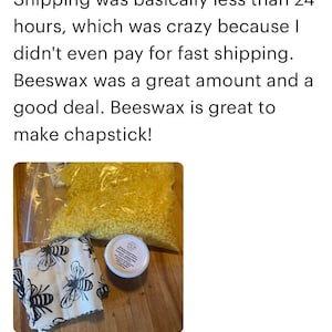 May include: A close-up of a bag of yellow beeswax pellets, a small white jar with a label, and a white fabric with a black bee pattern. The items are on a wooden surface. Beeswax Cosmetic Gr...