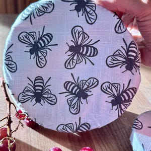 May include: A round fabric bowl cover featuring a repeating pattern of black bees on a white background. The circular textile cover is designed to fit over containers, showcasing a detailed insect print with visible fabric texture and stitching.