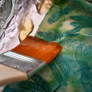 May include: A close-up of a paintbrush being used to apply a sealant to a fabric with a dragonfly pattern. The fabric is lying on a sheet of aluminum foil.