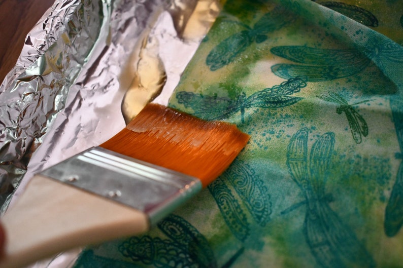 May include: A close-up of a paintbrush being used to apply a sealant to a fabric with a dragonfly pattern. The fabric is lying on a sheet of aluminum foil.