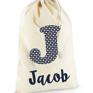 May include: A white canvas drawstring bag with a navy blue letter J with white stars on it. The name Jacob is written in navy blue cursive below the letter.