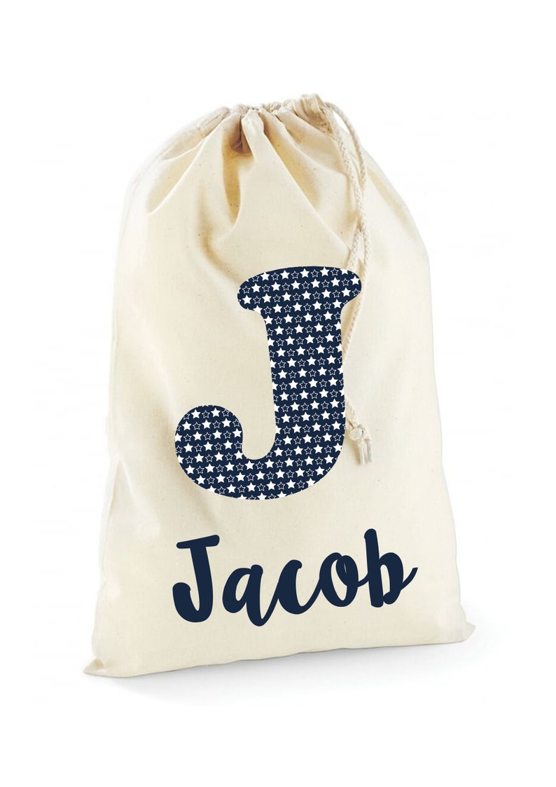 May include: A white canvas drawstring bag with a navy blue letter J with white stars on it. The name Jacob is written in navy blue cursive below the letter.