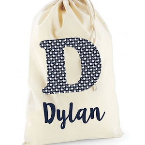 May include: A white canvas drawstring bag with a navy blue letter "D" printed on the front. The letter is filled with a pattern of white stars on a navy blue background. The name "Dylan" is printed below the letter in navy blue.