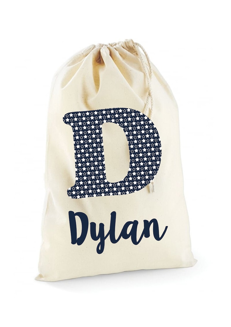 May include: A white canvas drawstring bag with a navy blue letter "D" printed on the front. The letter is filled with a pattern of white stars on a navy blue background. The name "Dylan" is printed below the letter in navy blue.