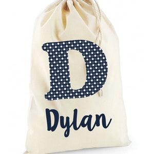 May include: A white canvas drawstring bag with a navy blue letter "D" printed on the front. The letter is filled with a white star pattern. The name "Dylan" is printed below the letter in navy blue.