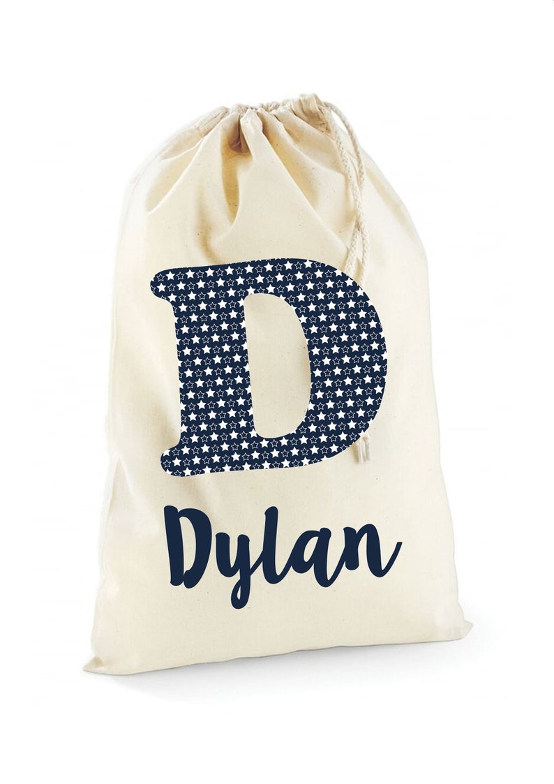 May include: A white canvas drawstring bag with a navy blue letter "D" printed on the front. The letter is filled with a white star pattern. The name "Dylan" is printed below the letter in navy blue.
