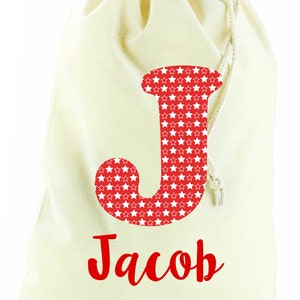 May include: A white canvas drawstring bag with a red letter "J" with white stars on it. The name "Jacob" is written in red cursive below the letter.