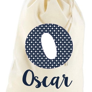 May include: A white canvas drawstring bag with a navy blue circle containing a white star pattern and the letter "O". The name "Oscar" is written in navy blue cursive below the circle.