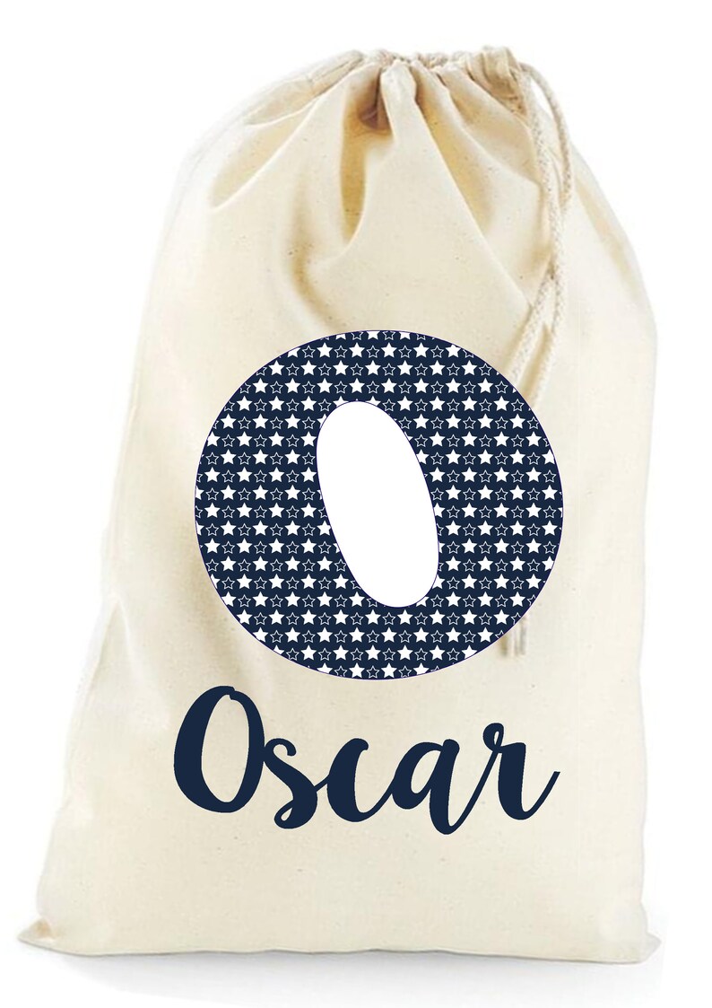 May include: A white canvas drawstring bag with a navy blue circle containing a white star pattern and the letter "O". The name "Oscar" is written in navy blue cursive below the circle.