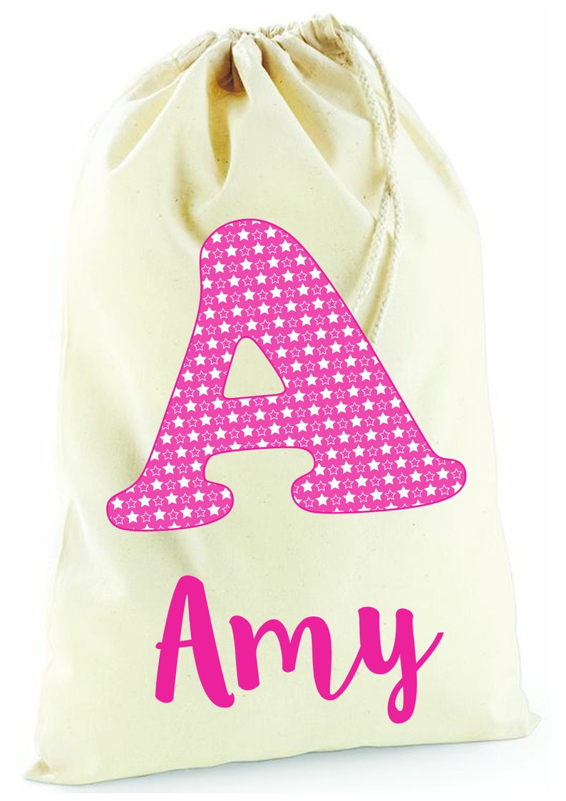 May include: A white canvas drawstring bag with a pink letter A covered in white stars and the name Amy written in pink below.