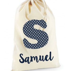 May include: A white canvas drawstring bag with a navy blue letter "S" printed on the front. The letter "S" is filled with a pattern of white stars on a navy blue background. The name "Samuel" is printed below the letter in navy blue cursive.