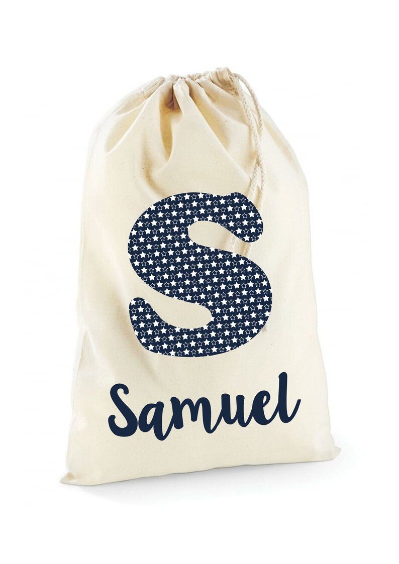 May include: A white canvas drawstring bag with a navy blue letter "S" printed on the front. The letter "S" is filled with a pattern of white stars on a navy blue background. The name "Samuel" is printed below the letter in navy blue cursive.