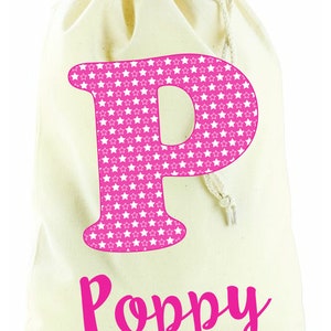 May include: A white canvas drawstring bag with a pink letter "P" printed on the front. The letter is filled with a pattern of white stars on a pink background. The name "Poppy" is printed below the letter in pink.
