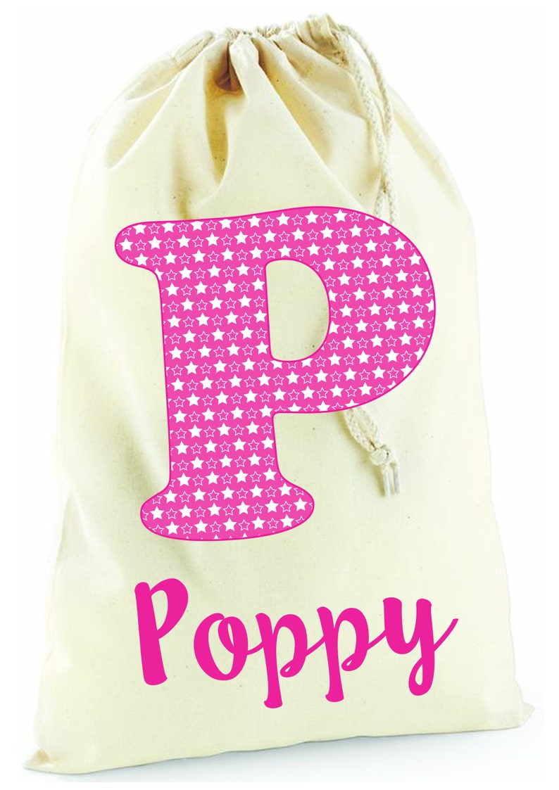 May include: A white canvas drawstring bag with a pink letter "P" printed on the front. The letter is filled with a pattern of white stars on a pink background. The name "Poppy" is printed below the letter in pink.