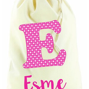 May include: A white canvas drawstring bag with a pink letter "E" covered in white stars and the name "Esme" written in pink cursive.