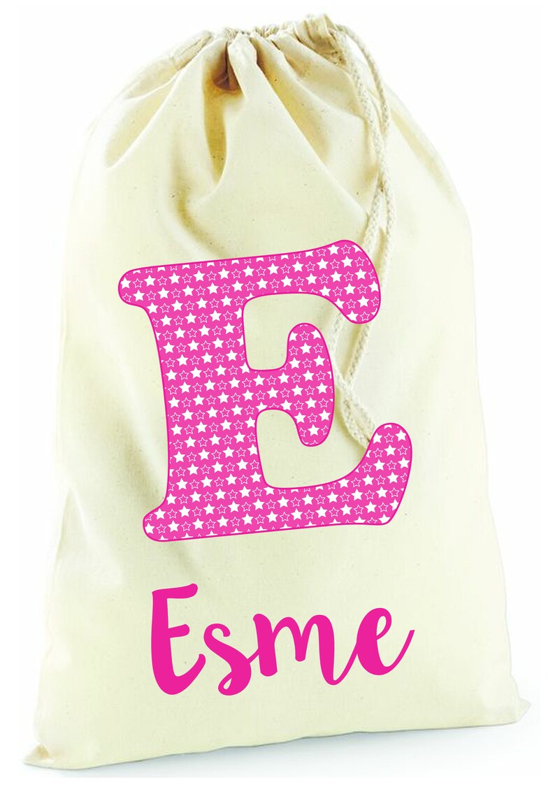 May include: A white canvas drawstring bag with a pink letter "E" covered in white stars and the name "Esme" written in pink cursive.