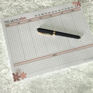 Könnte beinhalten: Ein Wochenplaner-Block mit dem Text "THIS WEEKS plan" in handschriftlichem Stil. Der Block hat ein Rasterlayout mit Wochentagen und Monaten. Ein schwarzer Stift mit goldenen Akzenten liegt auf dem Block.
