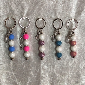 May include: Five beaded keychains with silver-tone rings. Each keychain features a different color scheme: blue and white, pink and white, purple and white, teal and white, and pink and brown. The beads are round and strung together.
