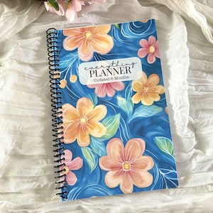 May include: A spiral-bound planner with a floral design. The cover features a blue background with orange and pink flowers and green leaves. The text "everything PLANNER" is displayed on a white label, with "Undated 6-Months" below.