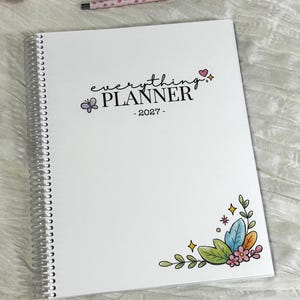 May include: A white spiral-bound planner with the text everything PLANNER - 2027 - printed on the cover. The cover features a small purple butterfly, a pink heart, and a colorful floral illustration in the bottom right corner with a matching pen.