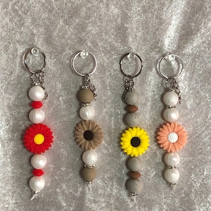 May include: Four keychains with flower and bead designs. Each keychain has a silver ring and chain, with a combination of white beads, flower-shaped charms, and other decorative beads in various colors like red, brown, yellow, and peach.