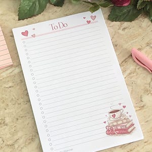Pode incluir: Um bloco de notas branco com linhas rosa e uma borda rosa. O bloco de notas tem as palavras "To Do" impressas no topo e uma xícara de café rosa com uma pilha de livros e corações na parte inferior. O bloco de notas está sobre uma superfície de mármore.