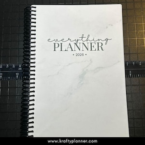 May include: A spiral-bound planner with a white marble-patterned cover. The text "everything PLANNER • 2026 •" is printed in gray script. The planner is open, and the black spiral binding is visible. The website address www.kraftyplanner.com is at the bottom.