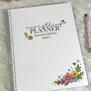 May include: A white spiral-bound planner with the text everything PLANNER Seasonal Edition - 2027 printed on the cover. The cover features colorful illustrations of flowers, leaves, small hearts, and stars in the top right and bottom right corners.