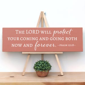 May include: A rectangular, coral-colored wooden sign with white lettering, displayed on a small wooden easel. The sign reads, "THE LORD WILL protect YOUR COMING AND GOING BOTH NOW AND forever. -PSALM 121:8-". A small potted plant sits below.