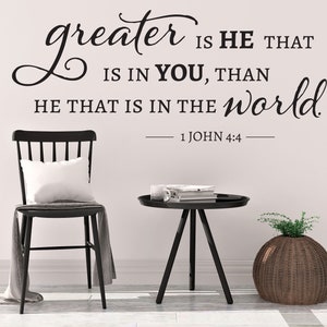 May include: Black and white wall decal with the quote "greater is HE THAT IS IN YOU, THAN HE THAT IS IN THE world."  The quote is from 1 John 4:4. The decal is displayed on a white wall in a room with a black chair, a black side table, and a wicker plant pot.