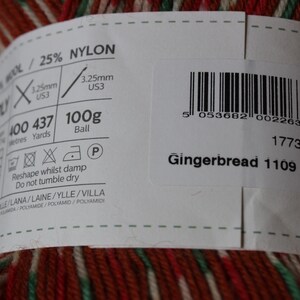 May include: A close-up of a ball of yarn with a label. The yarn is a mix of red, green, and white. The label reads "Gingerbread 1109" and includes details about the yarn's composition, weight (100g), and recommended needle size (US3).