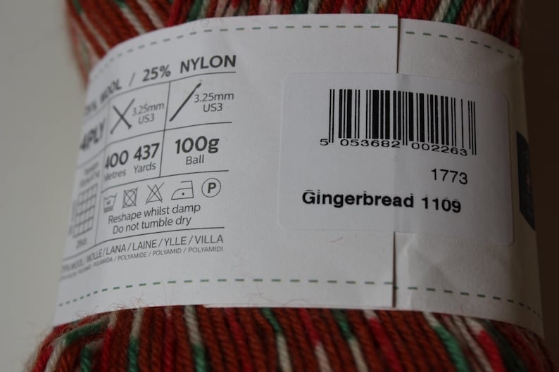 May include: A close-up of a ball of yarn with a label. The yarn is a mix of red, green, and white. The label reads "Gingerbread 1109" and includes details about the yarn's composition, weight (100g), and recommended needle size (US3).