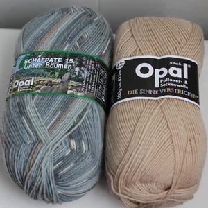 May include: Two skeins of Opal brand yarn. One is a mix of blue, gray, and orange, labeled "SCHAFPATE 15 Unter Bäumen." The other is a solid tan color. Both skeins have labels with the brand name and product details. Each skein weighs 100g.