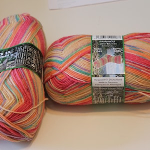 May include: Two skeins of Opal Schafpate 15 Unter Bäumen yarn featuring a variegated color palette of pink, orange, yellow, and teal. Each skein has a paper label with product information, a barcode, and an illustration of a finished sock.