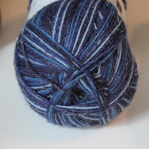 May include: A close-up of a ball of yarn in shades of blue, with silver metallic threads woven throughout. The yarn appears soft and textured, suitable for knitting or crochet projects. The color palette includes dark navy, light blue, and silver.