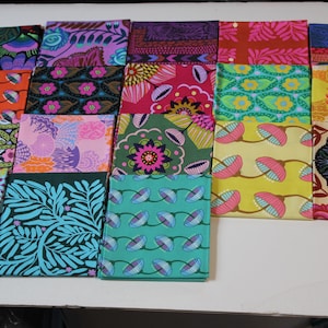 May include: A collection of folded fabric squares with vibrant floral and geometric patterns. The color palette includes shades of pink, blue, green, orange, and purple. The fabrics are arranged on a white surface.