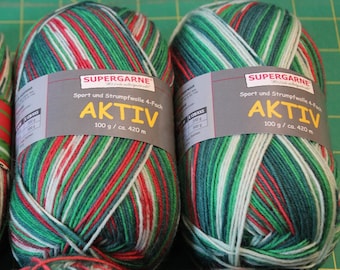 May include: Two skeins of Aktiv yarn with a mix of green, red, and white stripes. Each skein has a label that reads "SUPERGARNE AKTIV" and indicates 100 g / ca. 420 m. The yarn is designed for sport and sock knitting.
